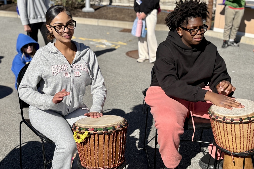 Two people play drums outdoors, seated on chairs in the sunlight.