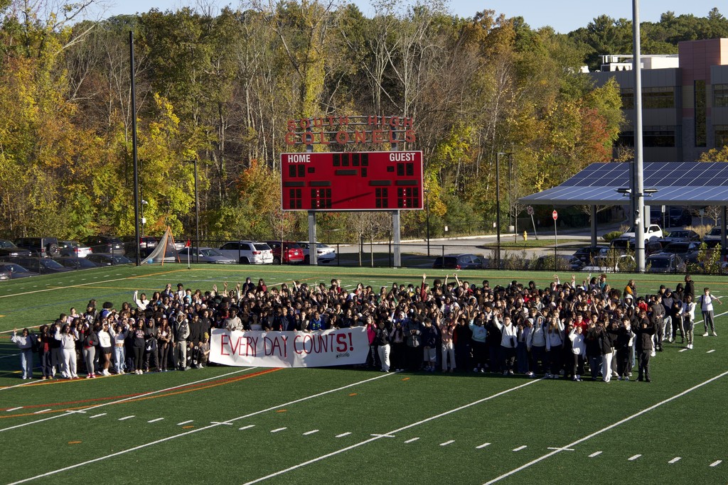 A large group of students stand on a football field, holding a banner.