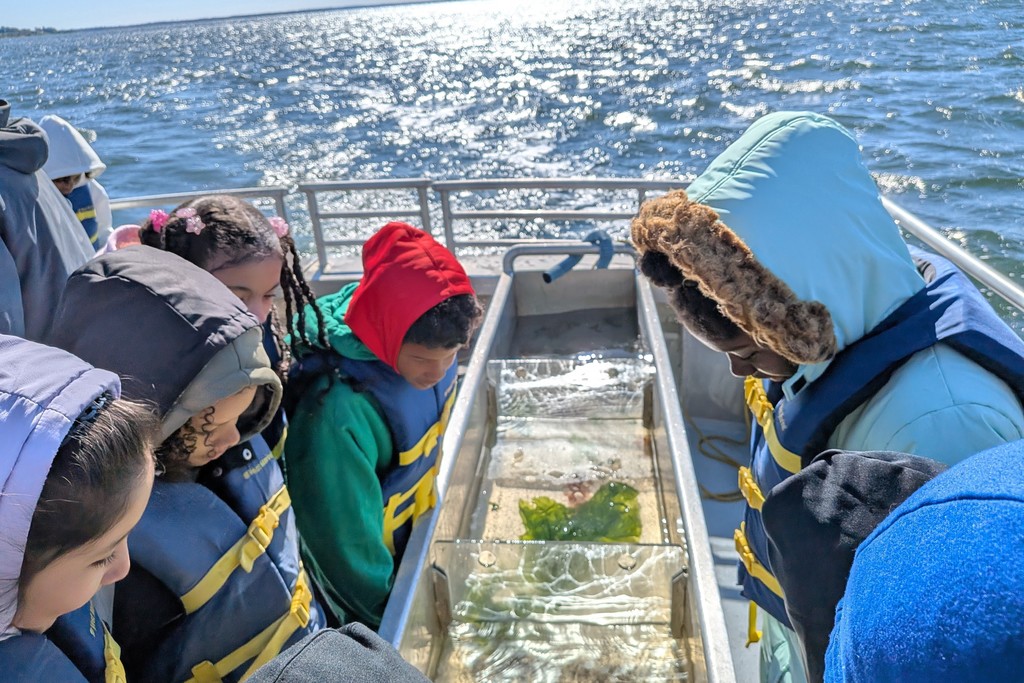 Children on a boat read informational pamphlets, wearing life vests.