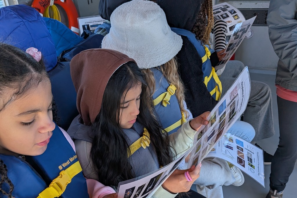 Children on a boat read informational pamphlets, wearing life vests.