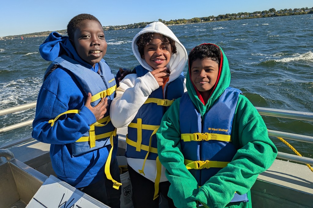 Three children wearing life vests smile on a boat, with water in the background.