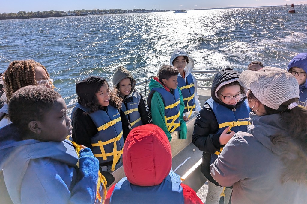 A group of children and adults wear life vests on a boat, enjoying a sunny day on the water.