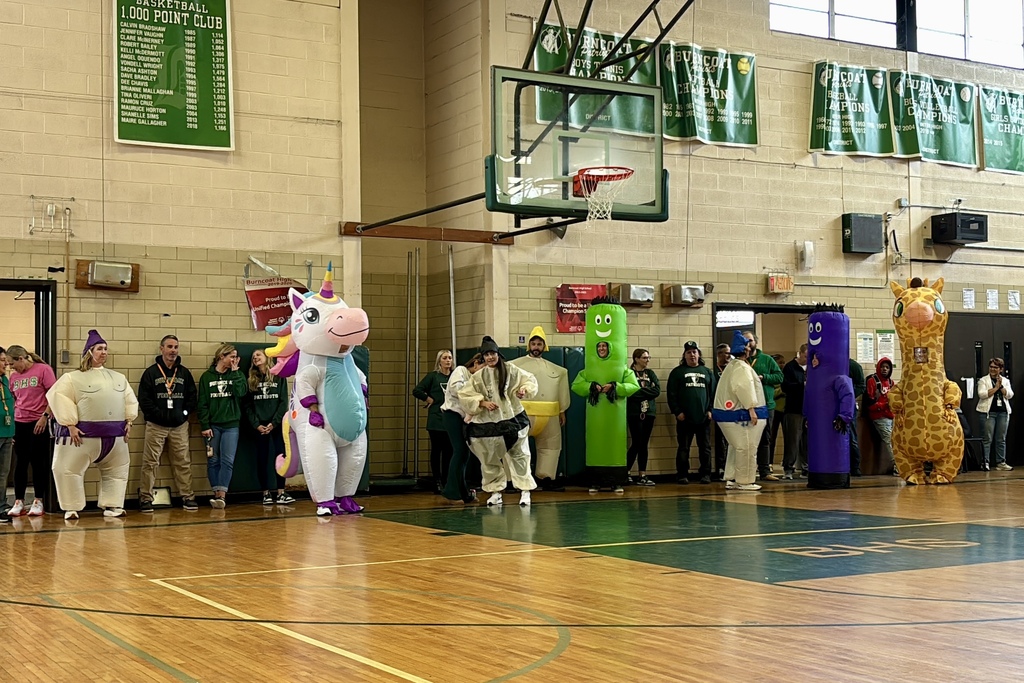 People in costumes stand on a basketball court, with banners and a scoreboard visible.