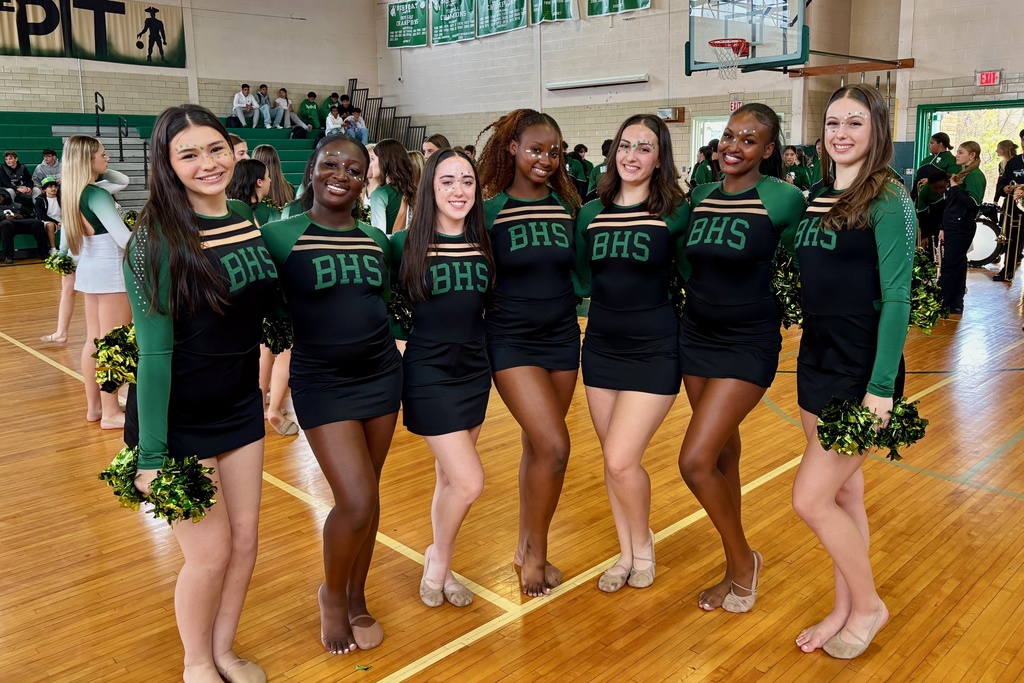A group of cheerleaders in black and green uniforms pose on a wooden basketball court.