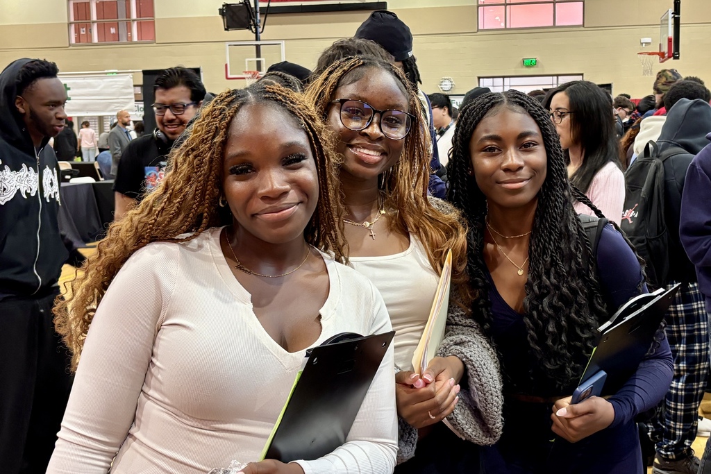 Three young women smile at the camera in a crowded indoor space, holding clipboards.