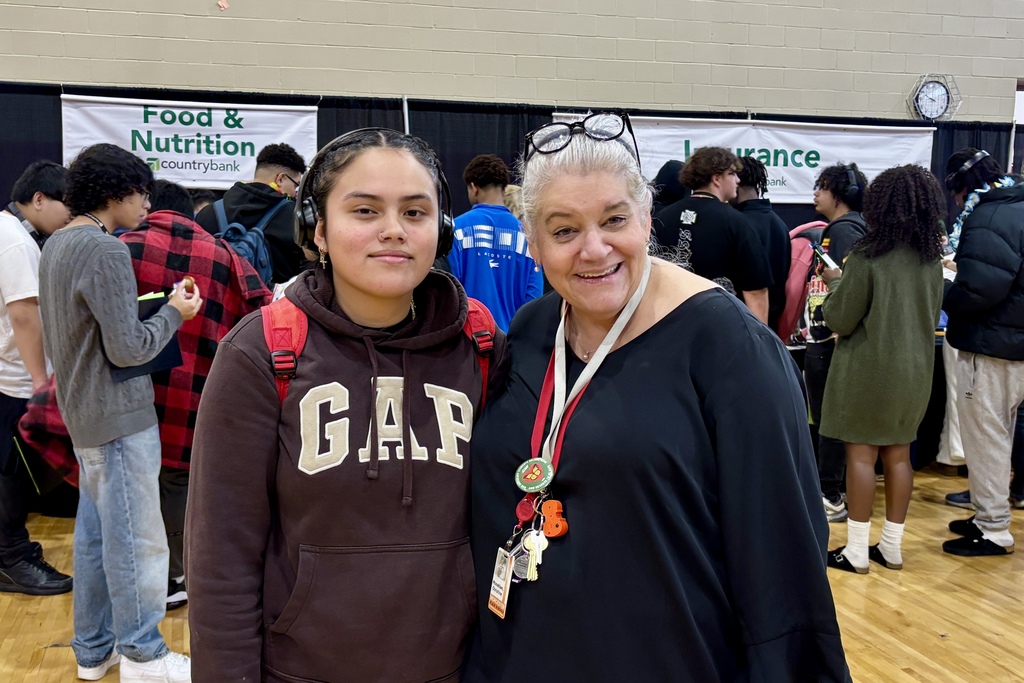 Two women smile at the camera in a crowded indoor space, possibly a school event.
