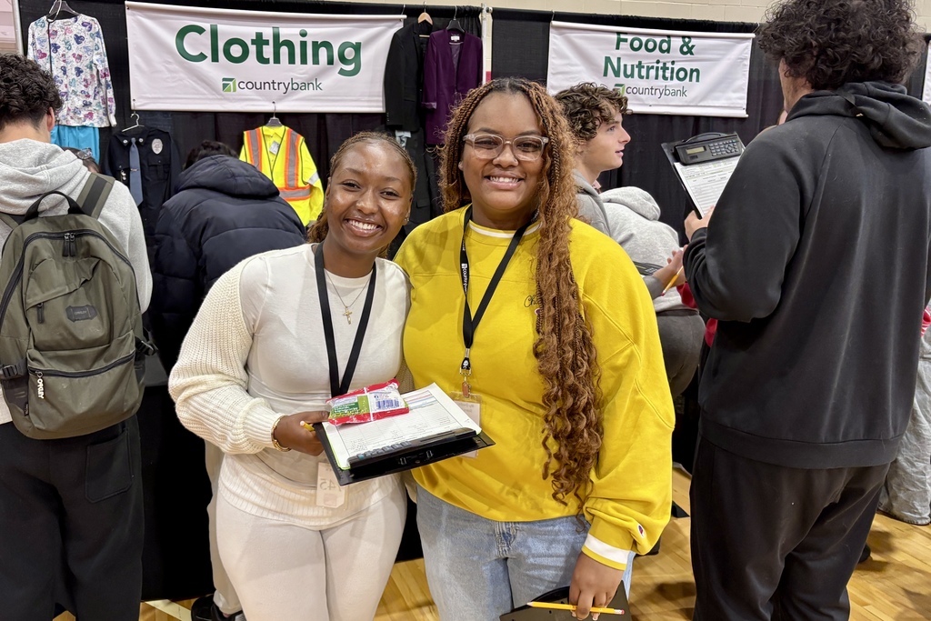 Two women smile at the camera at a trade show, holding clipboards.
