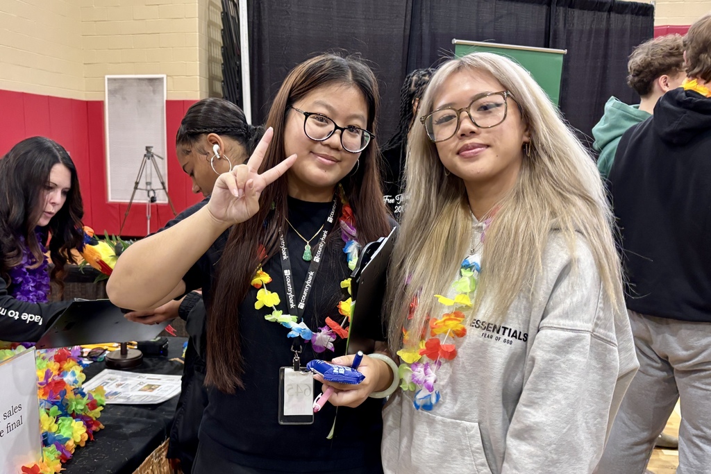 Two young women pose for a photo, one making a peace sign, both wearing leis.