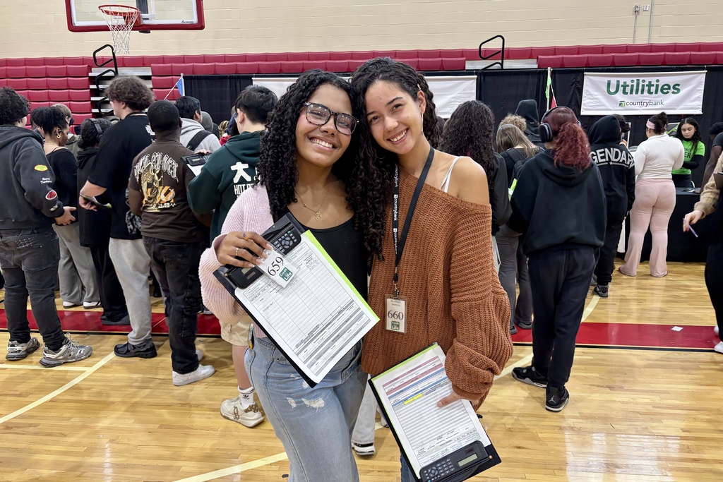 Two young women smile, holding clipboards in a crowded event space.