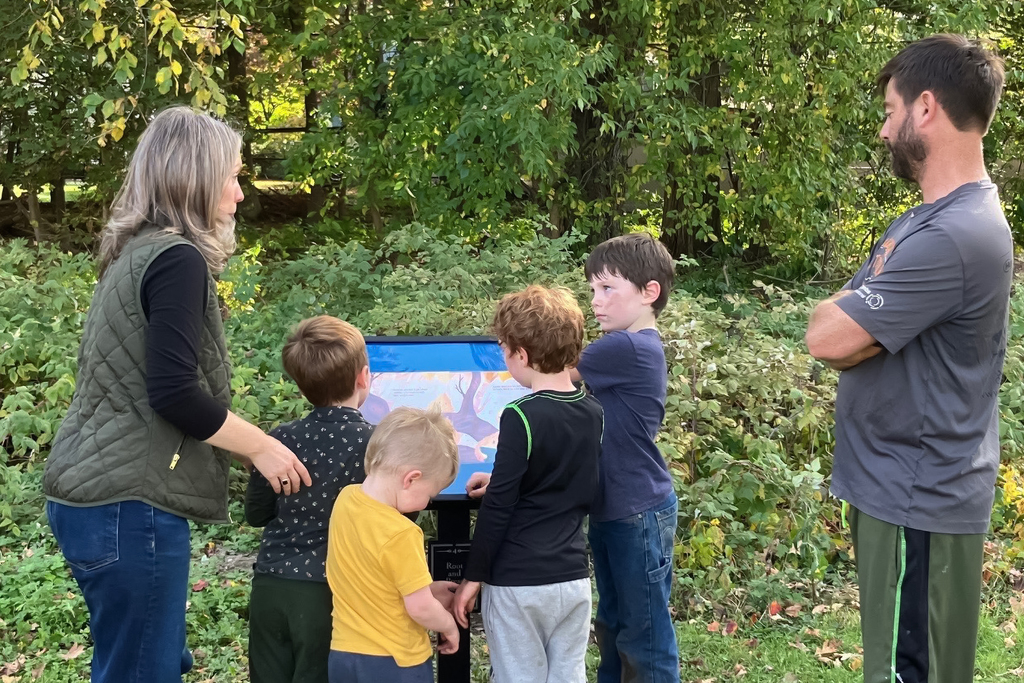 A group of people, including children and adults, gather around an informational sign outdoors.