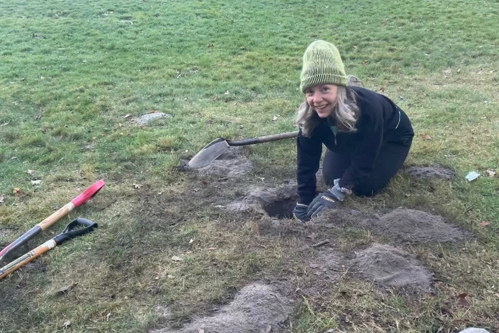 A woman kneels in a grassy area, digging a hole with a shovel.