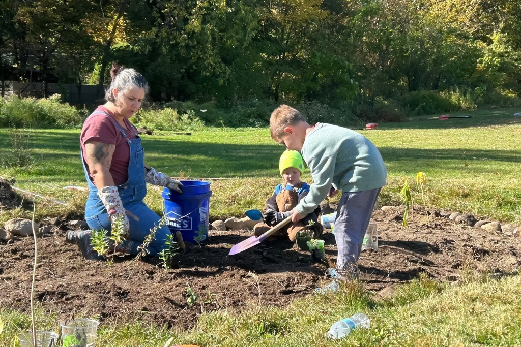 A woman and two children plant seedlings in a garden on a sunny day.