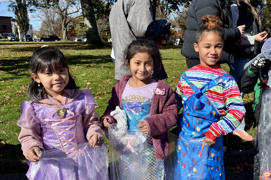 Three children in costumes smile for the camera outdoors on a sunny day.