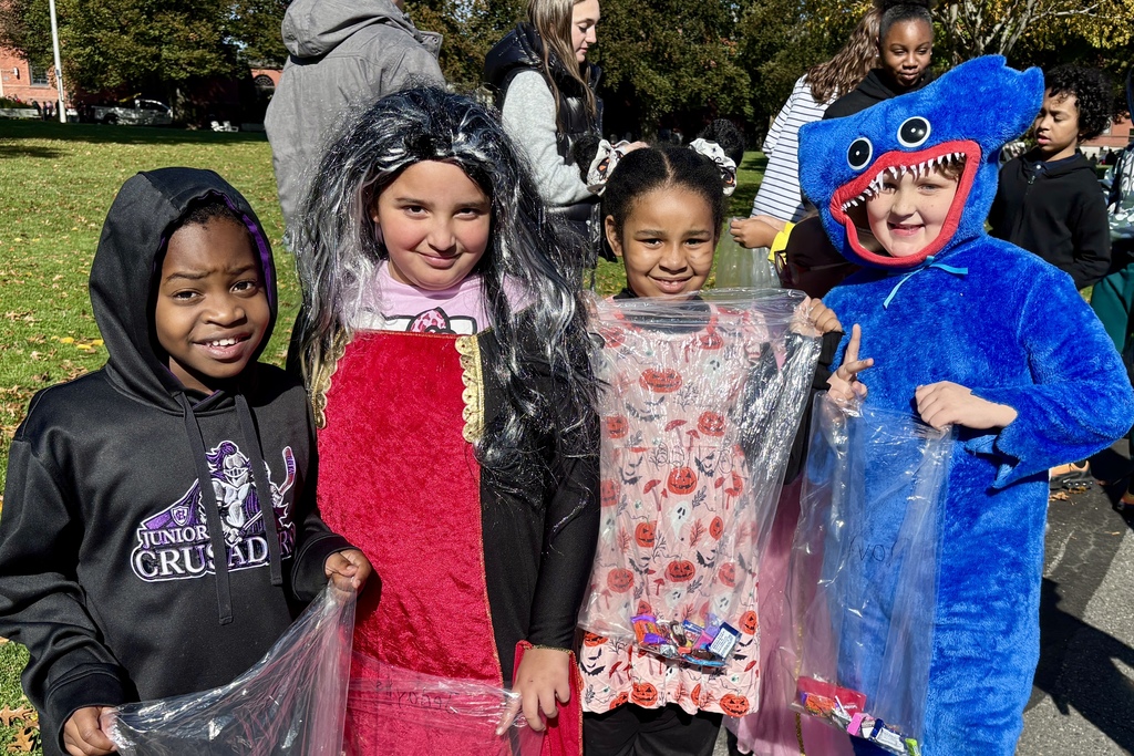 Children in Halloween costumes smile, holding treat bags outdoors on a sunny day.