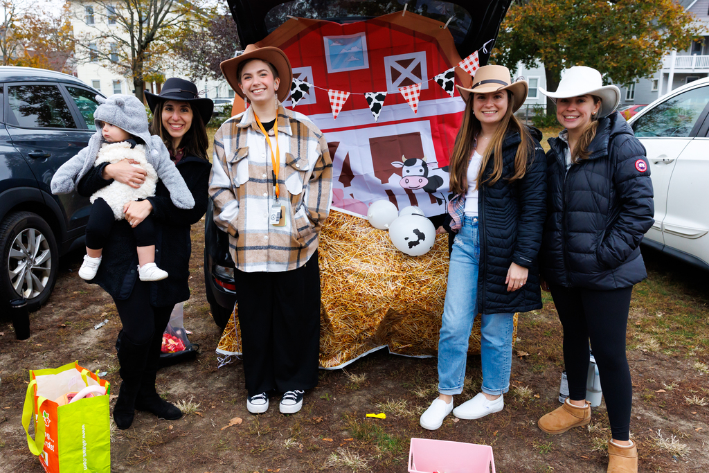 Four staff members and a baby, dressed as a sheep,  stand in front of their farmhouse display during a Trunk or Treat.