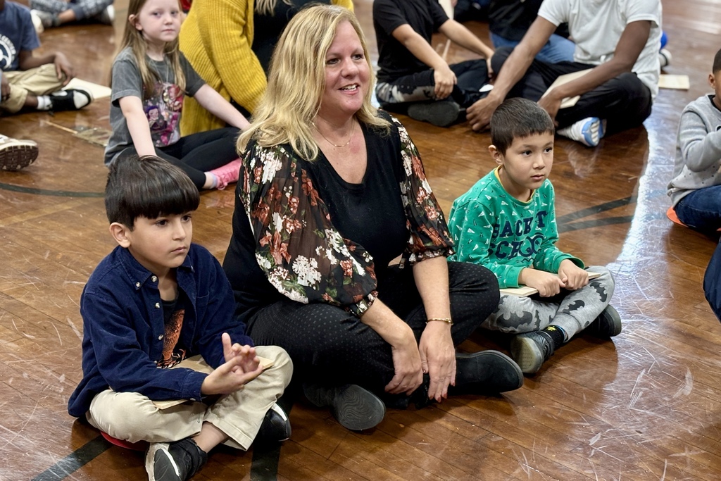 A group of children and an adult sit on a wooden floor, engaged in an activity.