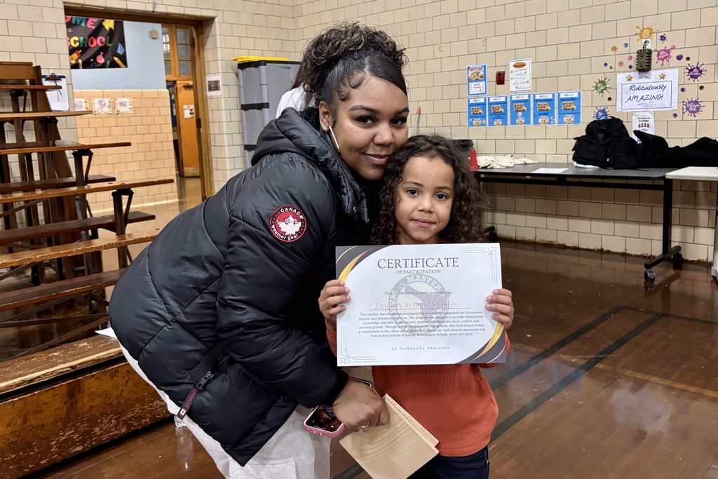 A woman and a young girl smile, holding a certificate in a school setting.