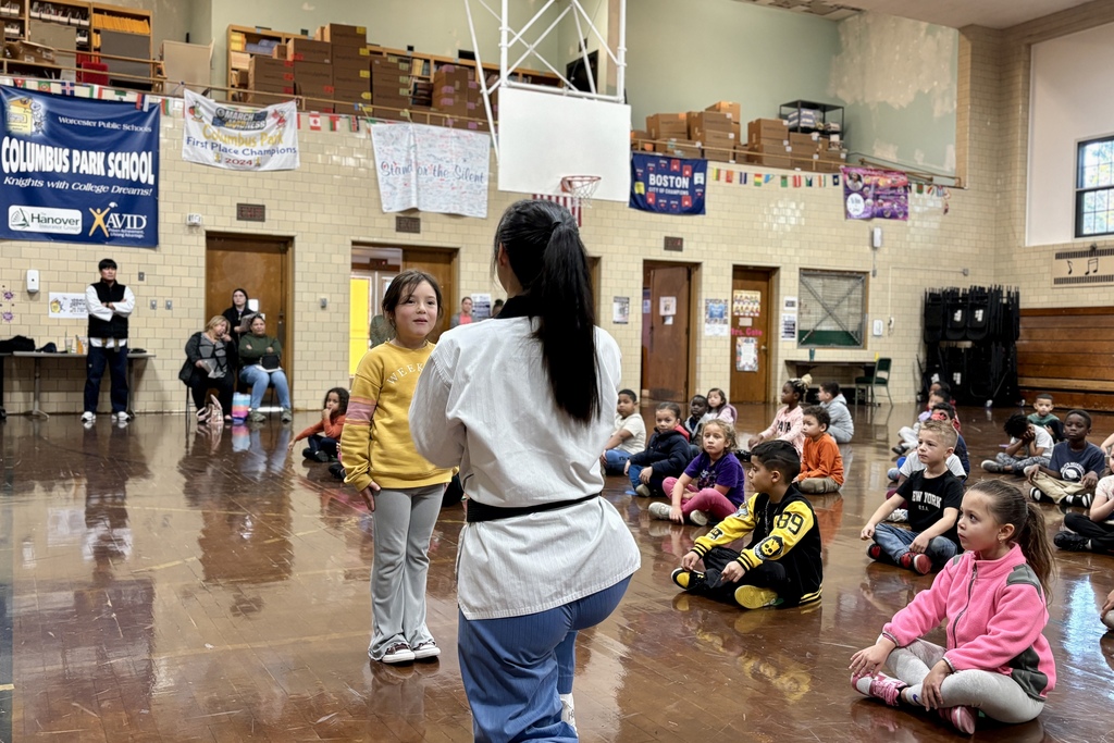 A martial arts instructor demonstrates a move to a young student in a school gymnasium.