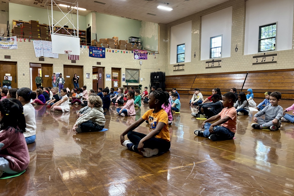 Children sit on the floor in a school gymnasium, facing forward.