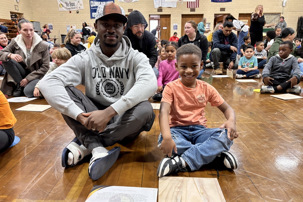 A man and a boy sit cross-legged on a wooden floor, smiling at the camera.