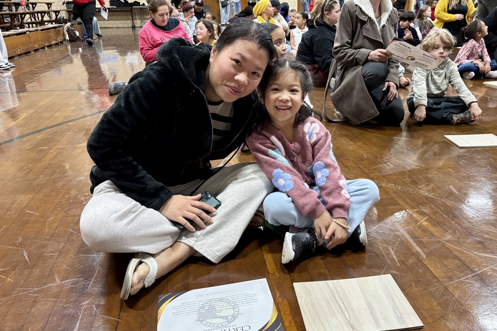 A woman and a young girl smile at the camera while sitting on a wooden floor.