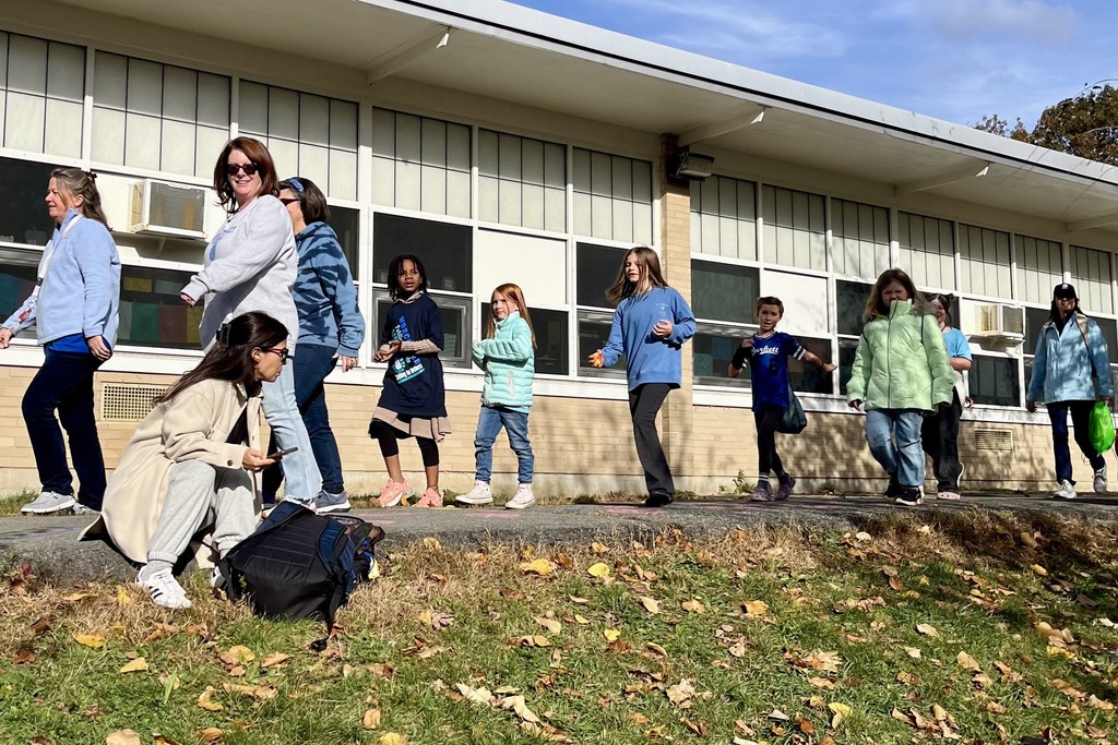 A group of people, including children and adults, walk along a sidewalk near a building.