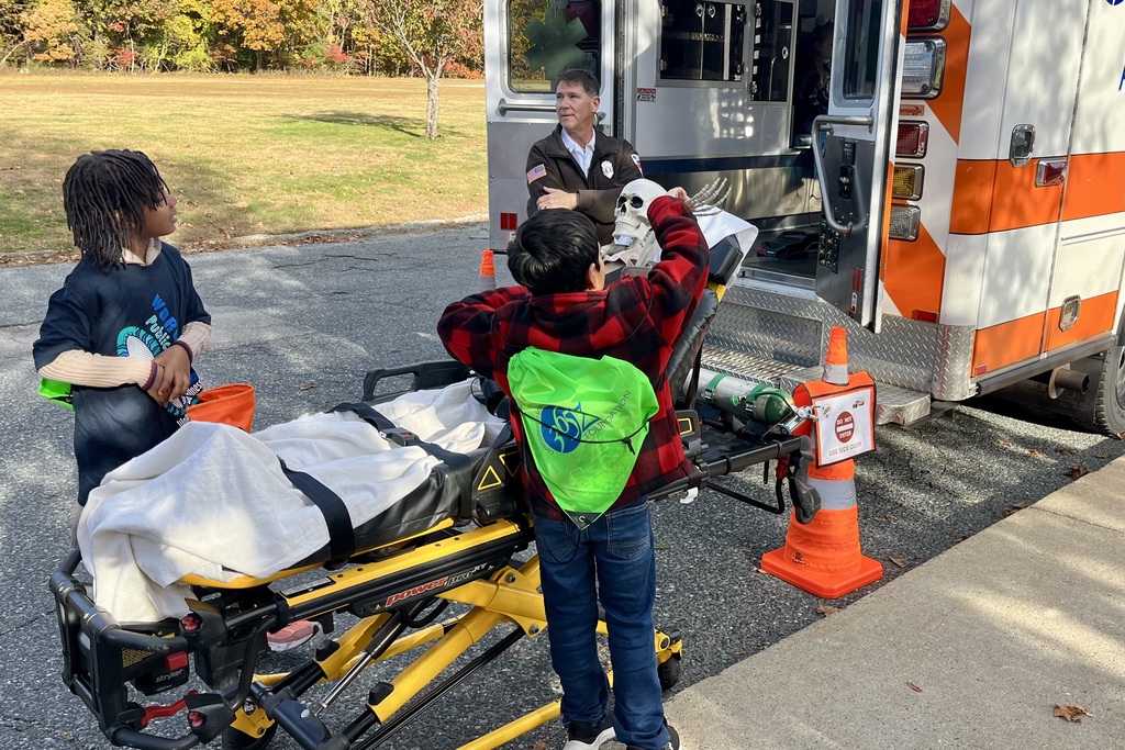 Two children and an adult stand near an ambulance, one child holding a skull.