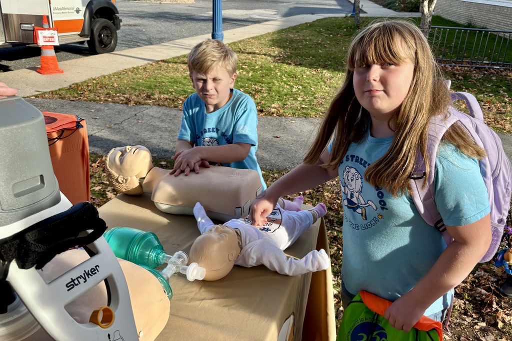 Two children practice CPR on mannequins outdoors, near a medical vehicle.