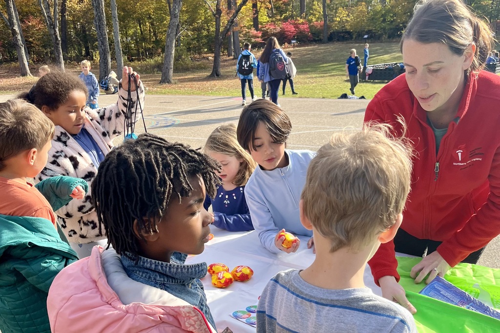 Children gather around a table with an adult, possibly for an outdoor activity.