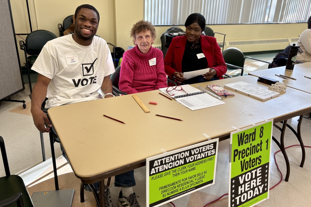 Three election officials sit at a table, smiling and ready to assist voters.