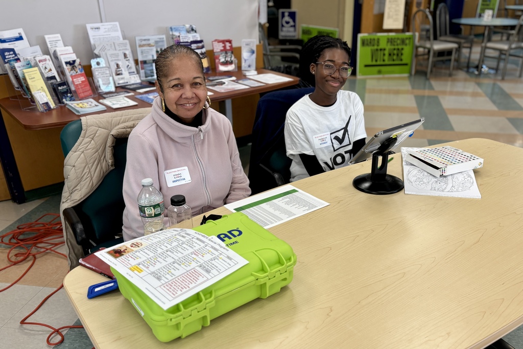 Two election officials sit at a table, smiling and ready to assist voters.