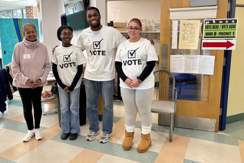 A group of people stand together, wearing 'VOTE' shirts, at a polling place.
