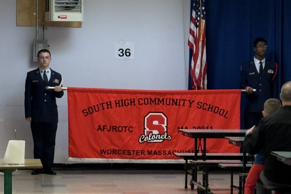 Two students in uniform stand holding flags in front of a banner.