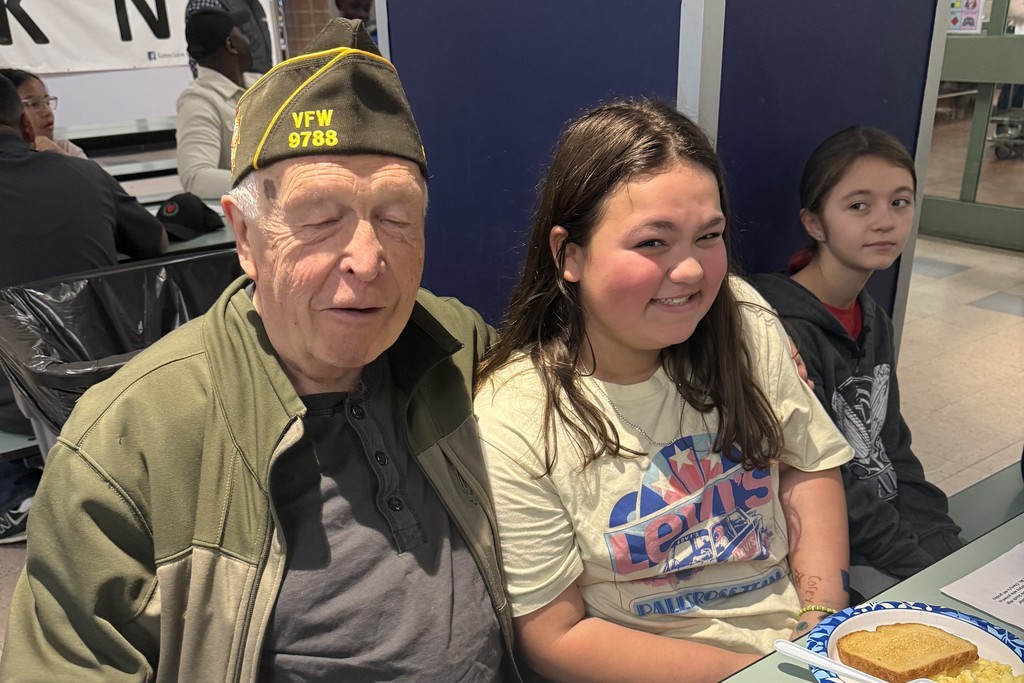 An older man in a VFW hat smiles next to a smiling young girl.