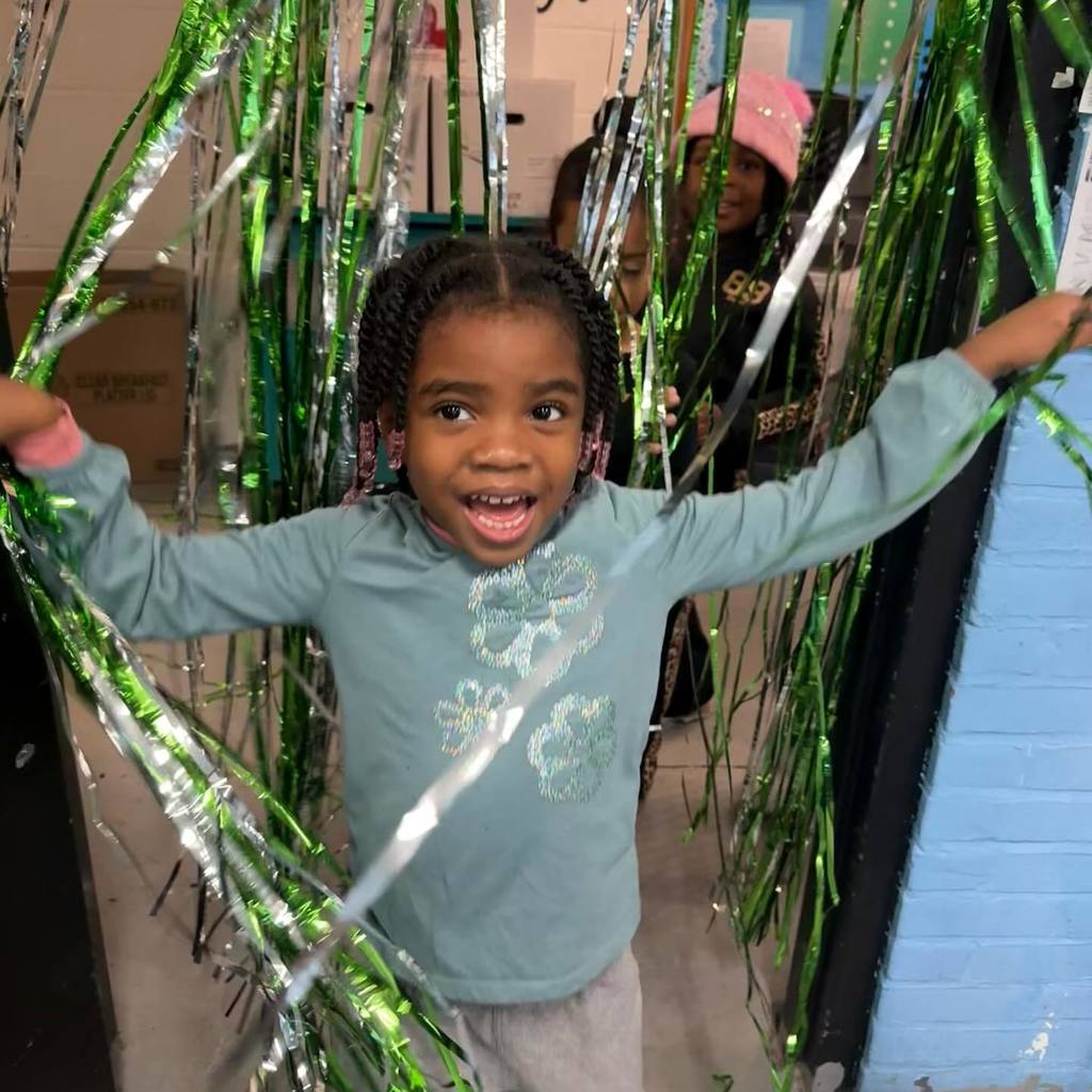 A young girl smiles with excitement, arms outstretched amidst green streamers.