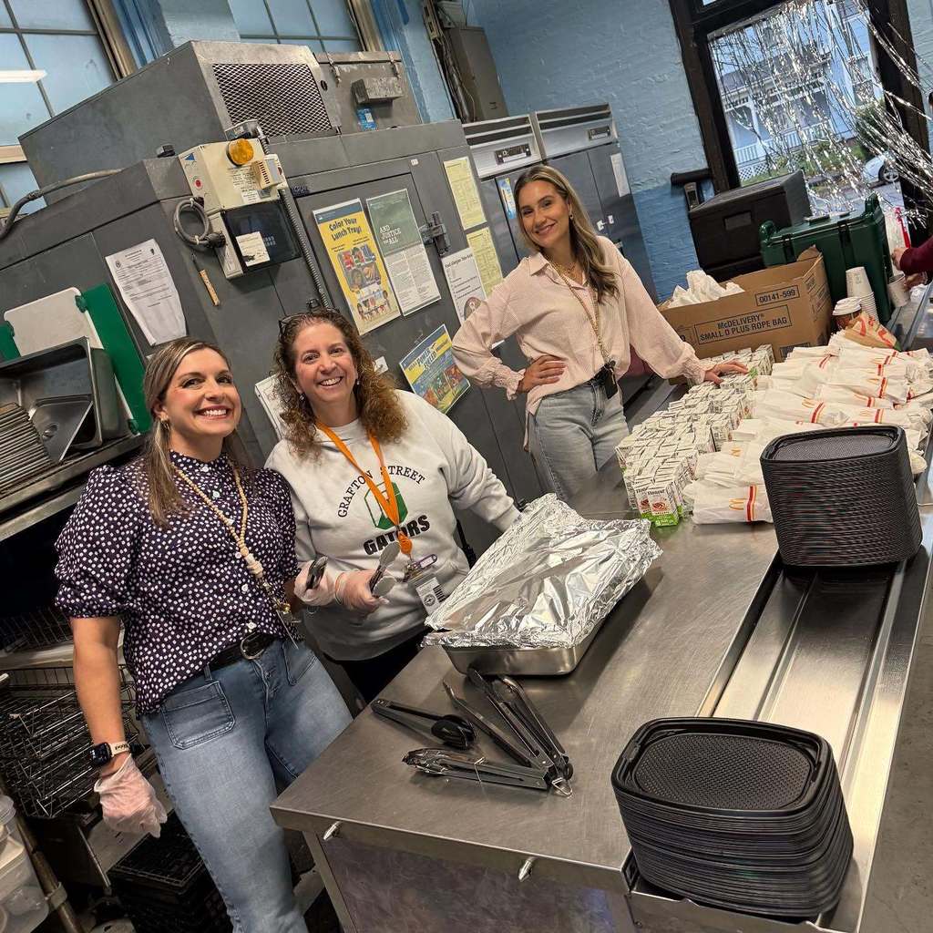 Three women smile in a commercial kitchen, preparing food.
