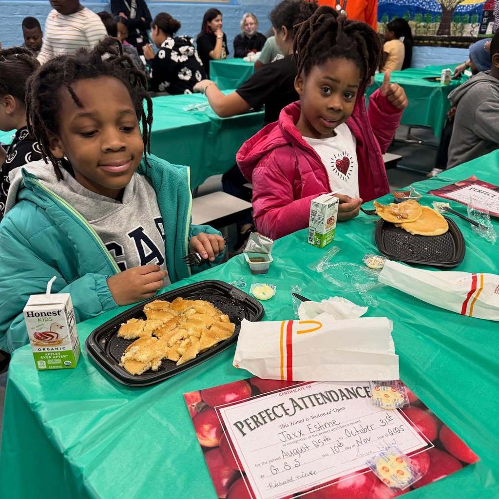 Two children enjoy a meal at a table, one giving a thumbs up.