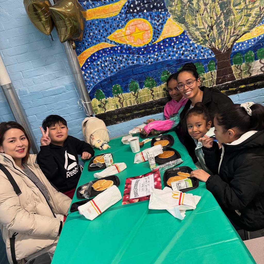 A group of people smile around a table with food and a colorful mural.