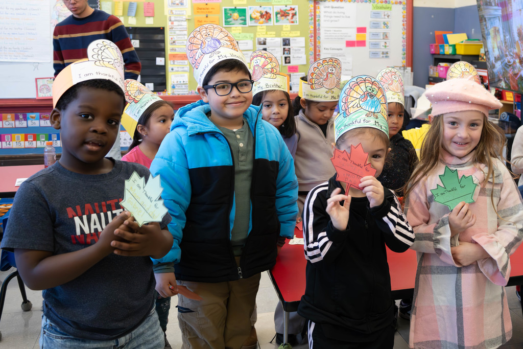 Children in a classroom wear turkey hats and hold leaf-shaped signs.