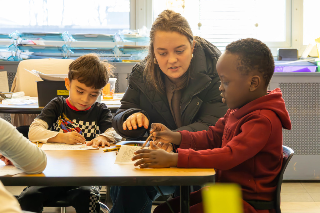 A teacher assists two students with a drawing activity at a classroom table.