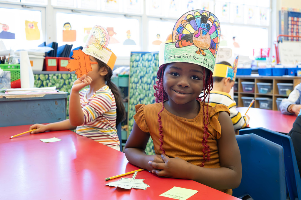 A young girl smiles at the camera, wearing a colorful turkey hat in a classroom.