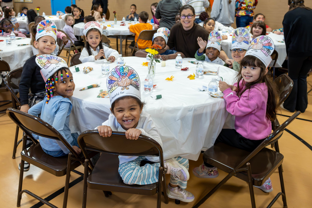 Children wearing turkey hats smile around a table, celebrating a holiday.