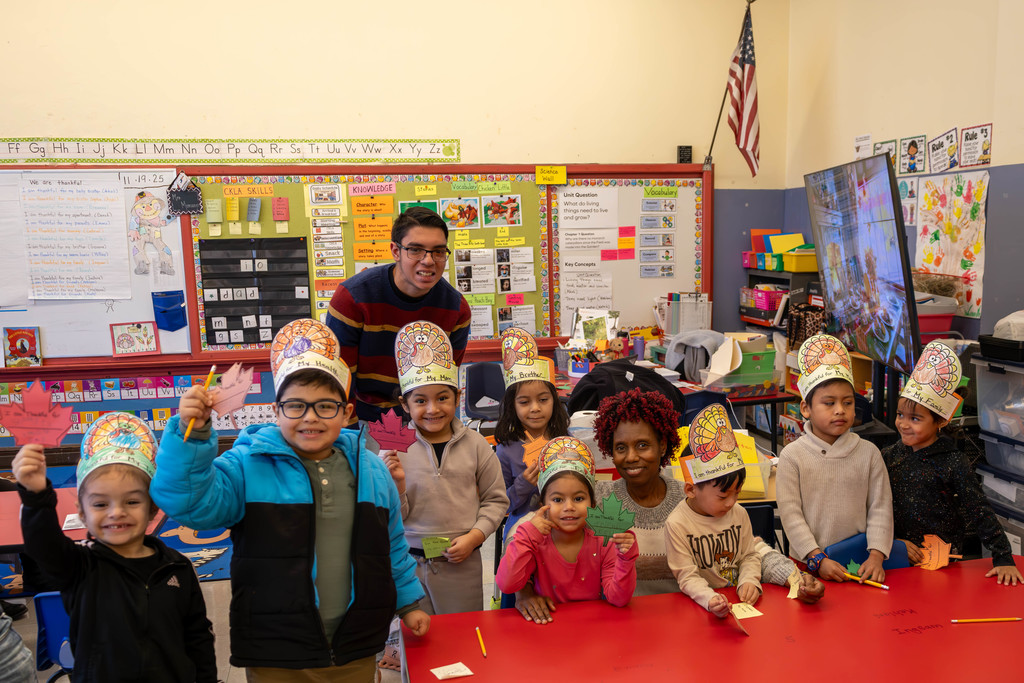 A classroom of children and adults celebrate Thanksgiving, wearing turkey hats and holding thankful notes.