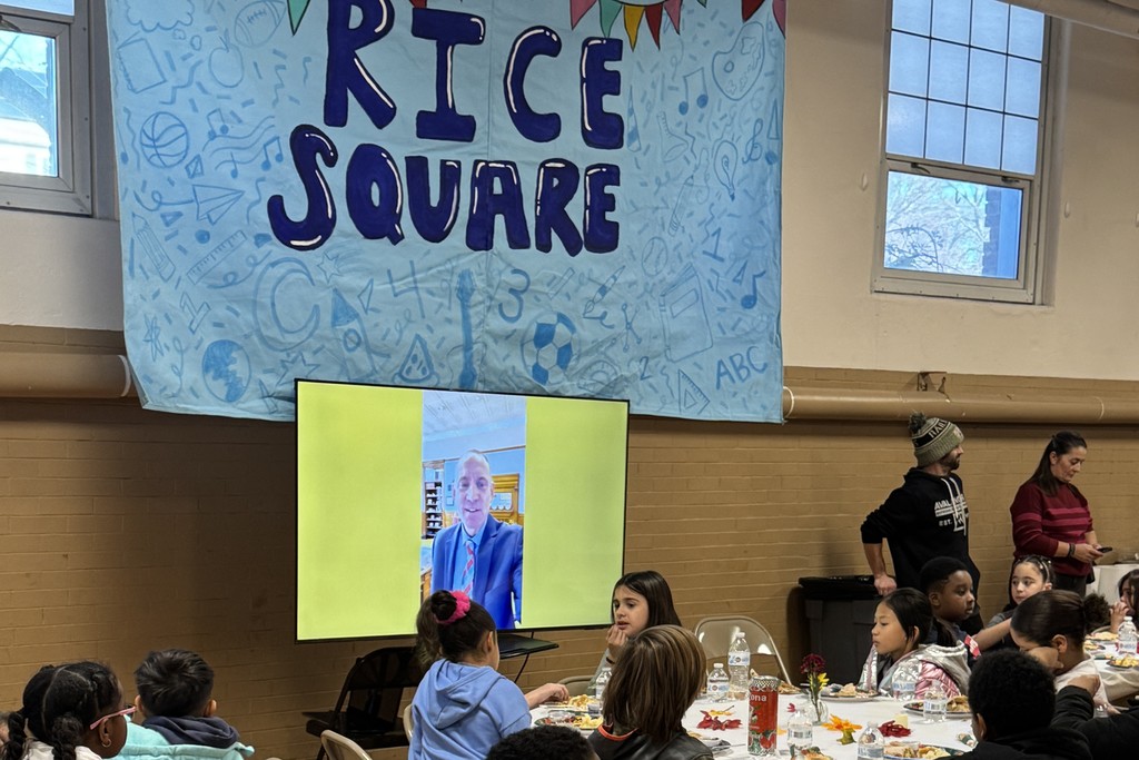 Children and adults gather at tables, watching a video call on a large screen.