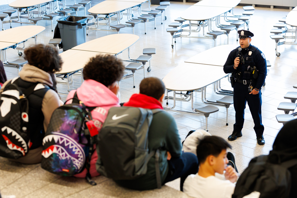 A police officer stands in the front of a group of students and speaks into a microphone during a Career Day presentation.