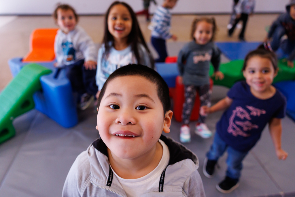 A  young child smiles at the camera. There are four other young students blurred in the background smiling.