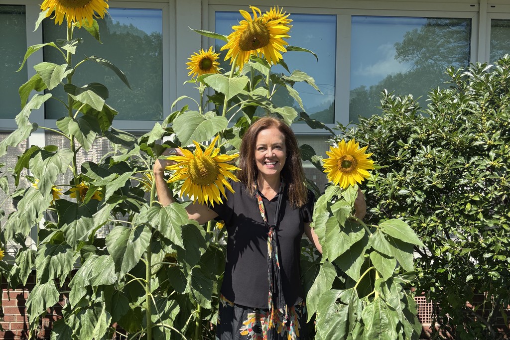 A woman smiles, standing among tall sunflowers in a garden.