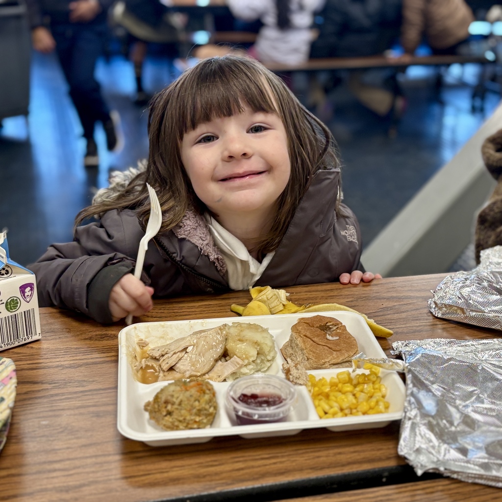 A young girl smiles at the camera while holding a fork, ready to eat her lunch.