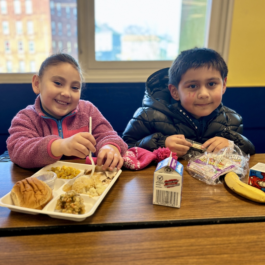 Two children smile while eating lunch at a table, with food trays and milk.