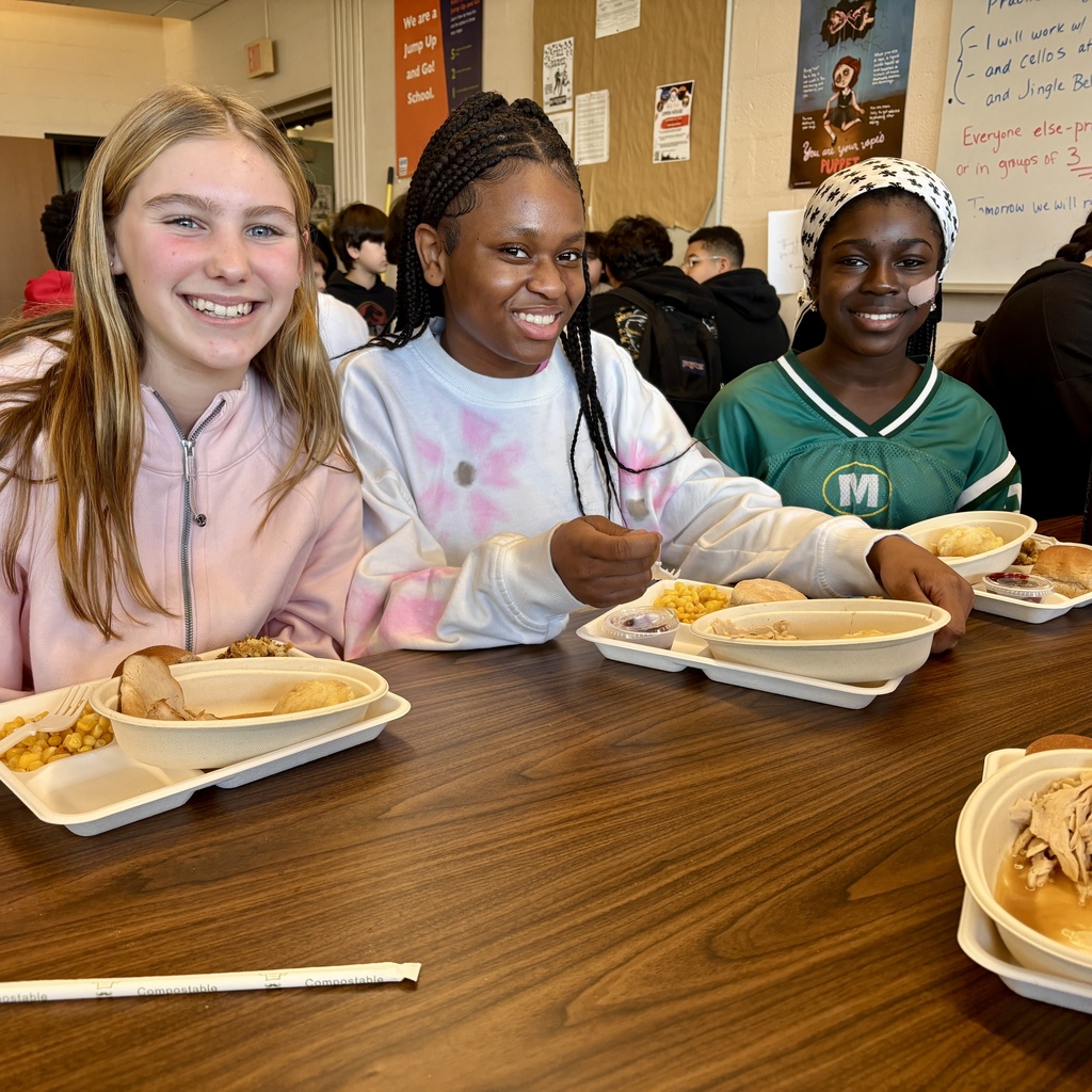 Three smiling students enjoy a meal at a school cafeteria table.